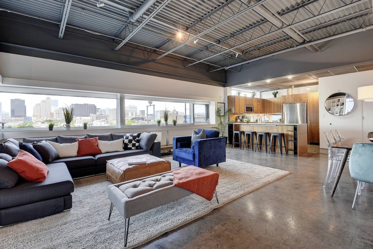 A spacious living area is highlighted by large windows offering city views. A sectional sofa in dark tones is accompanied by colorful accent pillows. A coffee table and two accent chairs provide additional seating. A modern kitchen with wooden cabinetry and a dining area are also visible.