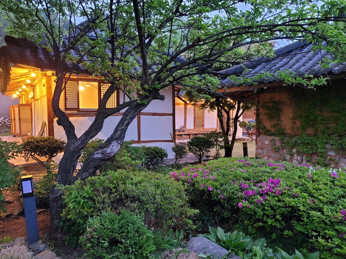 A traditional hanok is viewed through the branches of a tree, surrounded by lush greenery and blooming flowers. Soft lighting emanates from the structure, creating a serene atmosphere in the garden. The natural stone walls can be seen blending harmoniously with the landscape.