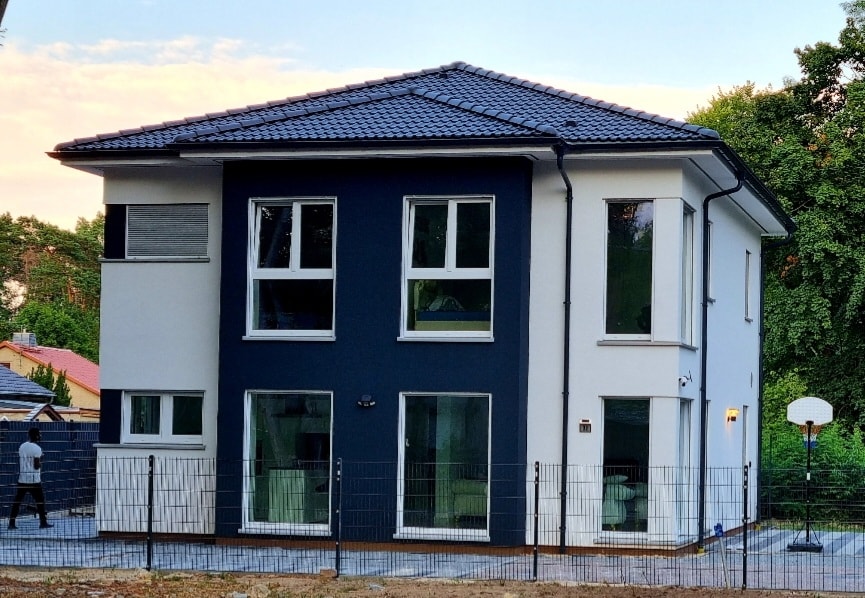 The modern two-story house features a combination of white and dark blue exteriors, highlighted by large windows that allow natural light. A fenced outdoor area is visible, providing a space for leisure. The surrounding green trees enhance the peaceful ambiance of the home.
