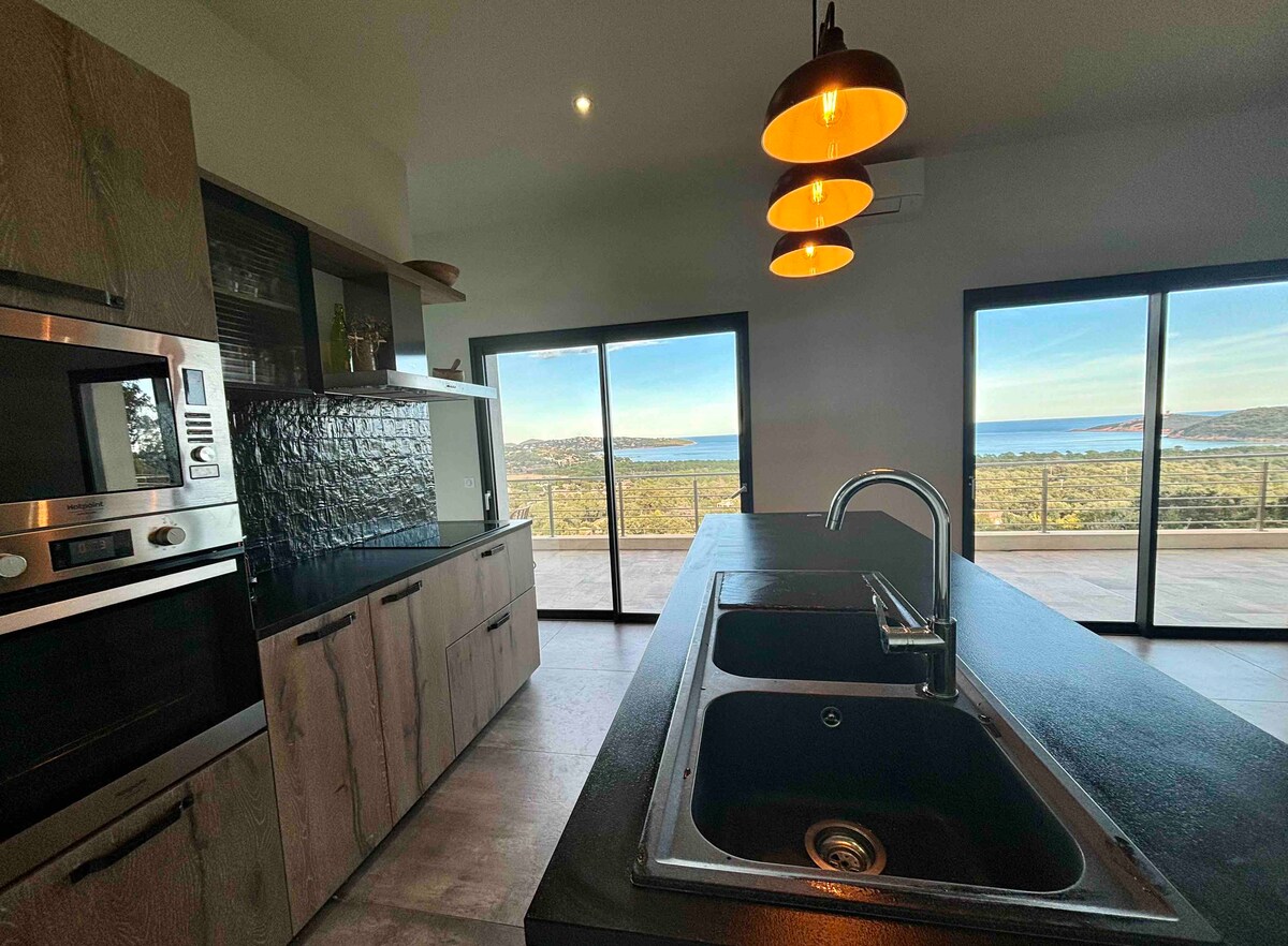 A modern kitchen is designed with sleek cabinetry and equipped with stainless steel appliances. A large island with a double sink is centered in the space. Floor-to-ceiling glass doors offer panoramic views of the surrounding landscape and water, bringing natural light into the area.