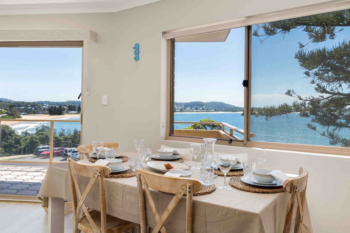 A dining area is presented with a light brown table set for six, adorned with glassware and woven placemats. Large windows showcase stunning ocean views, allowing natural light to illuminate the space and create a serene dining atmosphere.