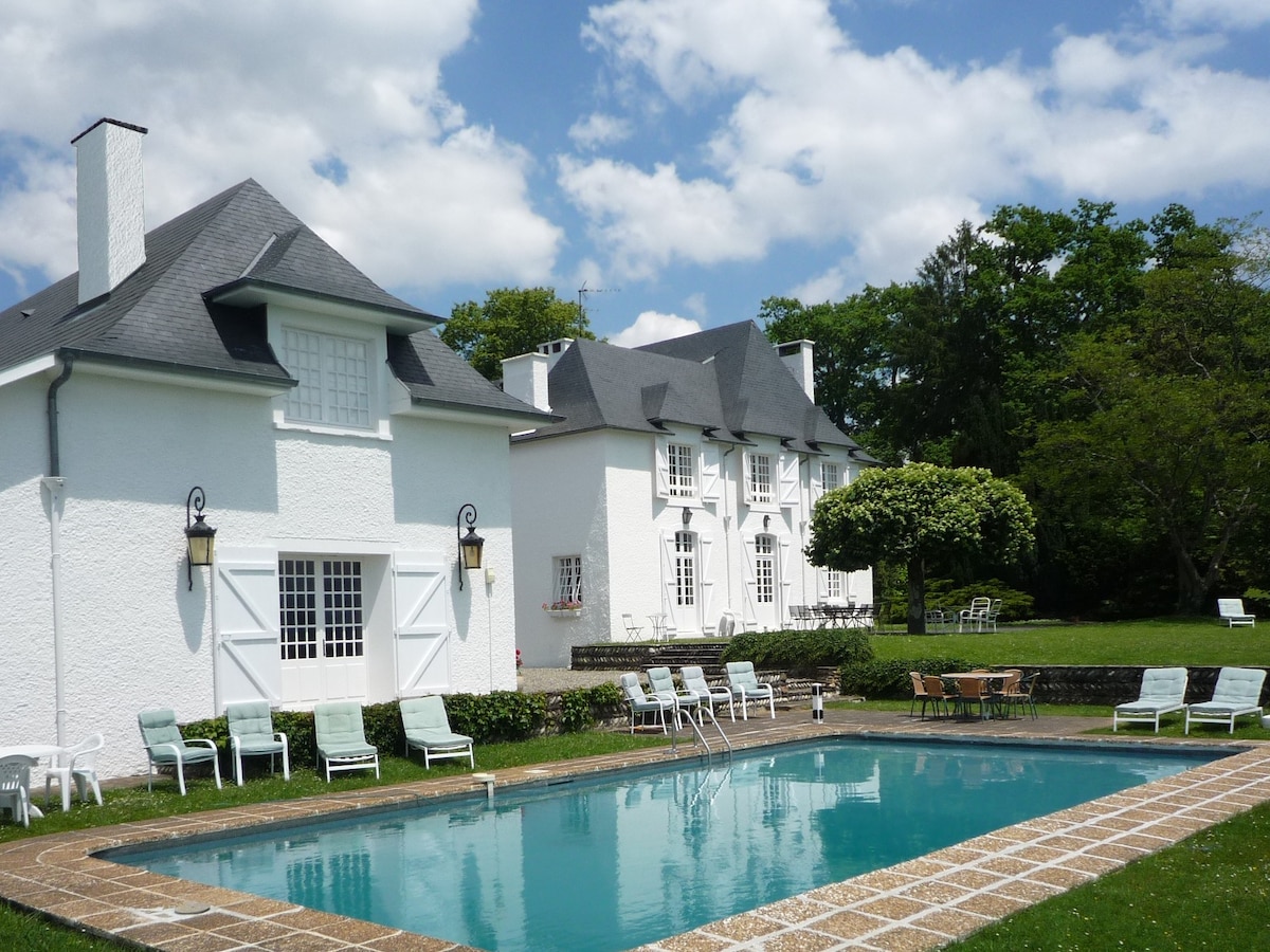 The image depicts a white manor house surrounded by lush greenery, showcasing a sparkling outdoor pool. Lounge chairs are arranged around the poolside, inviting relaxation. The sky is partially cloudy, adding a serene touch to the tranquil landscape.