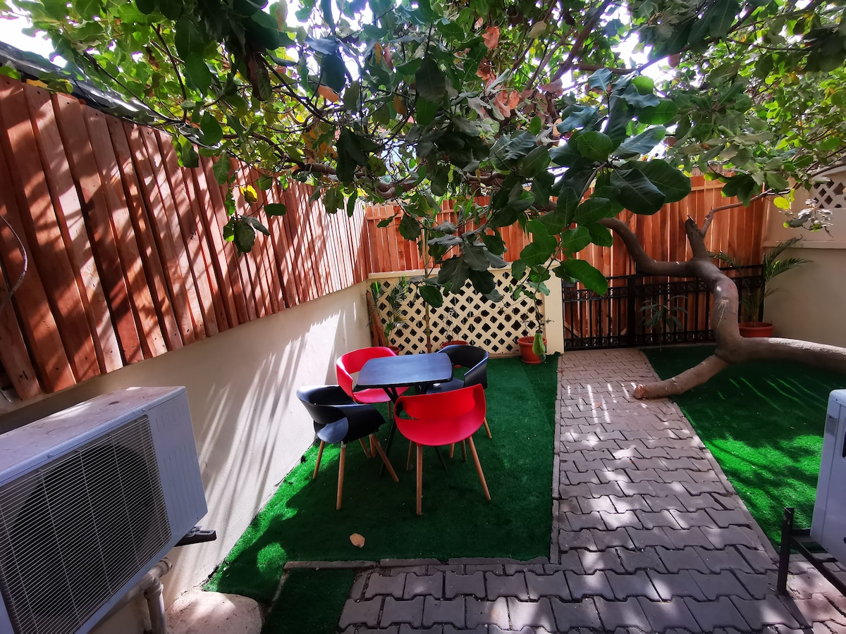 An outdoor dining area is set on a grassy surface, featuring a black table surrounded by four colorful chairs—two red and two black. The space is shaded by a leafy tree, with walls partially enclosed for privacy.