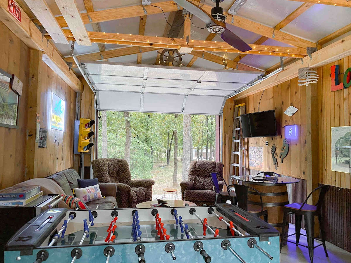 A game room features a foosball table in the foreground, with a cozy seating area behind it. Large doors open to a wooded view, allowing natural light to fill the space. Ambient lighting adds a relaxed atmosphere with a television mounted on the wall.