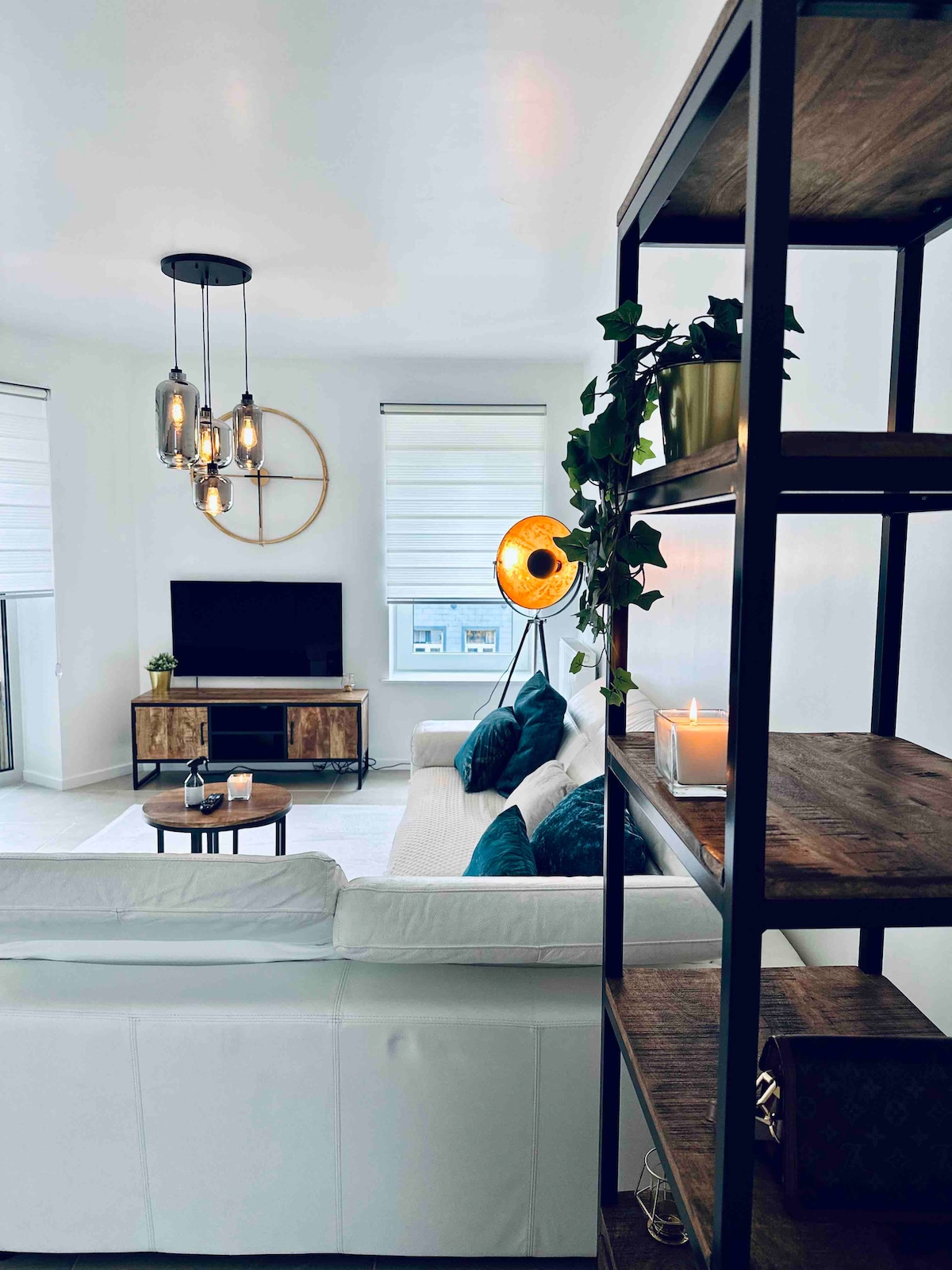 A modern living area features a comfortable light-colored sofa adorned with teal pillows, accompanied by a wooden coffee table. A flat-screen television is positioned on a wooden media unit, while large windows with roller blinds allow natural light to illuminate the space.