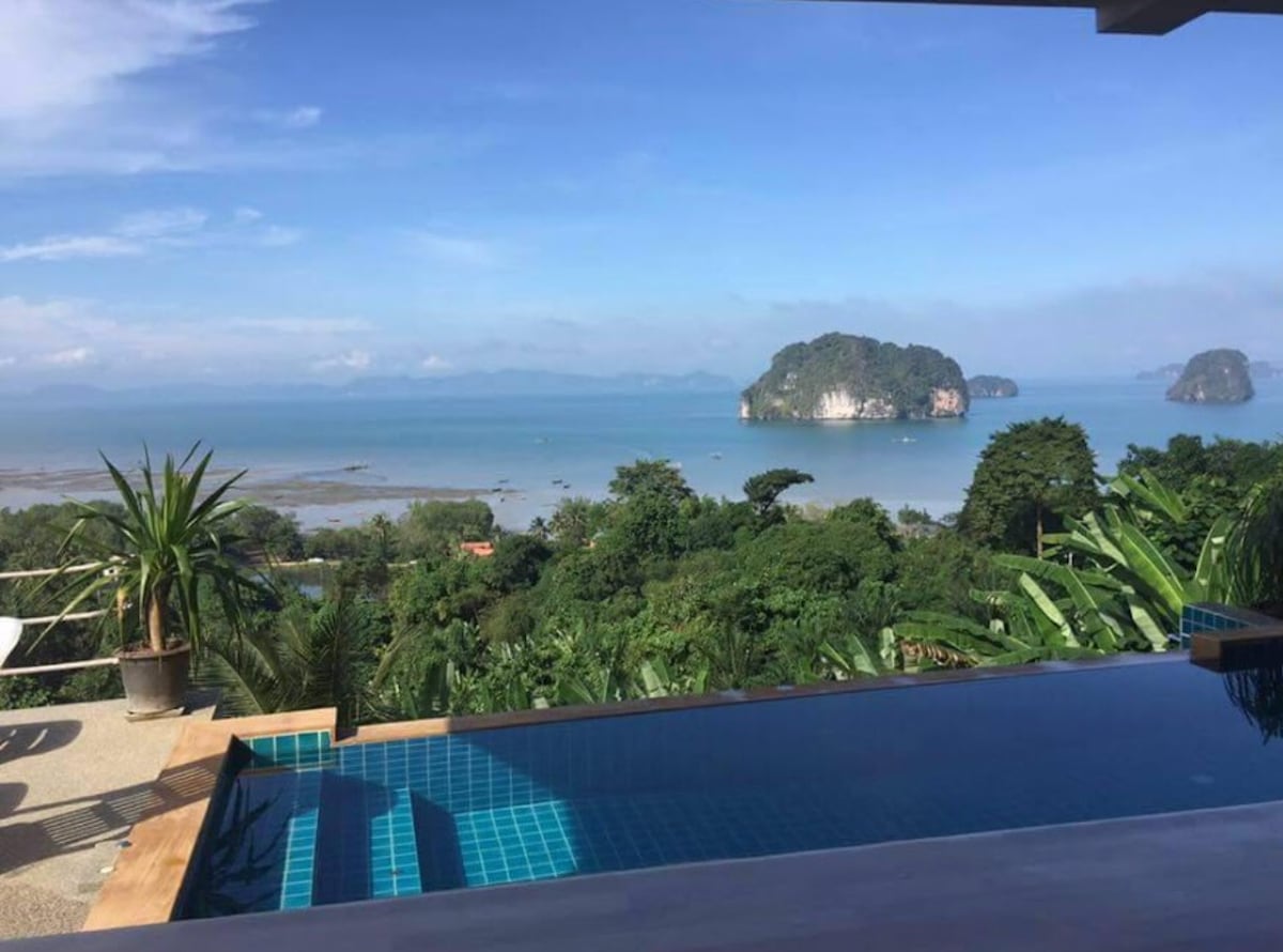 An expansive view of the ocean is captured from a poolside vantage point. Lush greenery surrounds the infinity pool, which reflects the clear blue sky and nearby rocky islands. The serene coastal landscape stretches into the distance, providing a sense of tranquility.