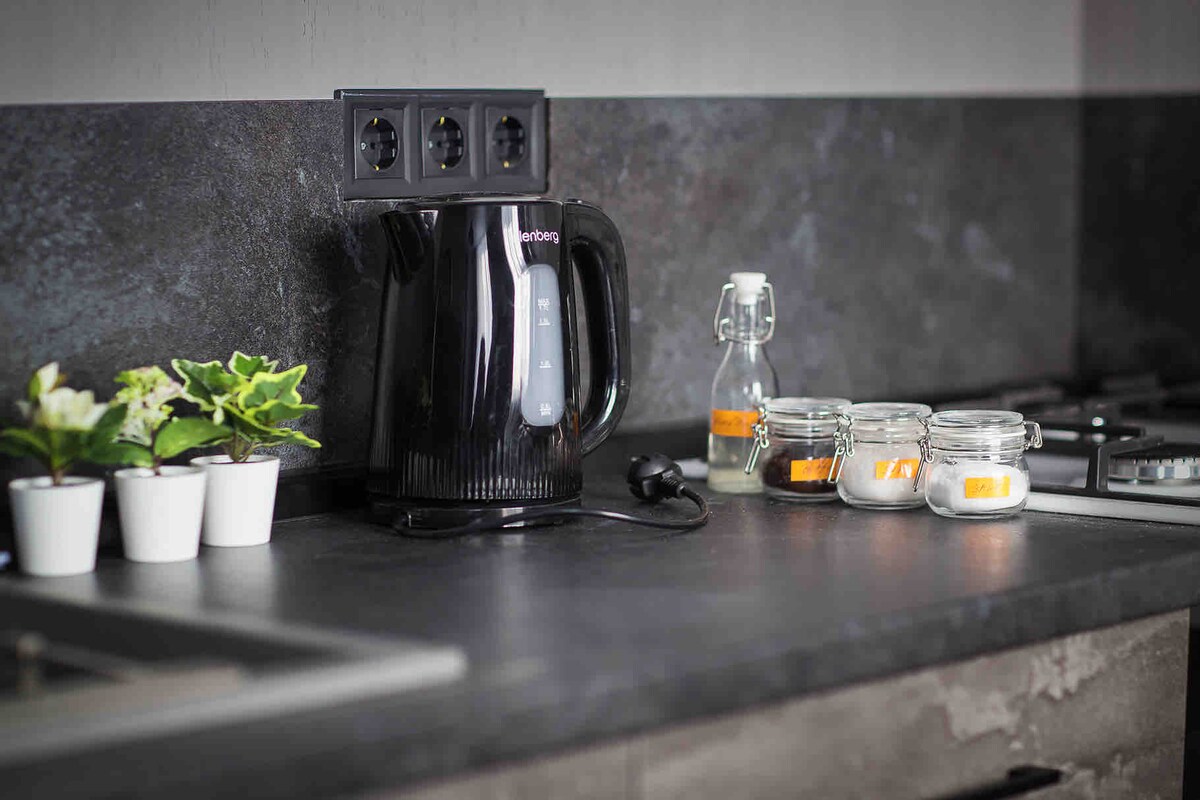 A modern kitchen counter features an electric kettle alongside glass jars filled with various ingredients. Small potted plants add a touch of greenery, while a clear glass bottle sits nearby. The countertop is finished in dark material, complementing the sleek design.