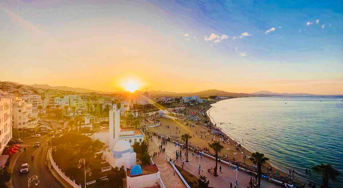 A sunset view captures the sun setting over the beach, with golden light reflecting on the water. Palm trees line the promenade, and people can be seen enjoying the shoreline. Nearby buildings create a lively urban backdrop against the serene coastal scenery.