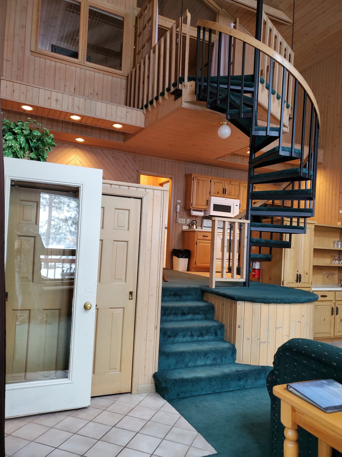A spiral staircase leads to an upper level of the chalet, surrounded by warm wooden walls. An open door reveals a closet, while a microwave and other kitchen amenities are visible in the background. Soft teal carpeting covers the staircase and landing area.