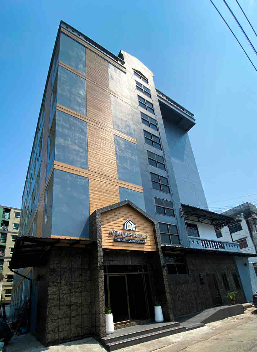 The exterior of a modern multi-story building showcases a blend of metal and wooden elements. Large windows reflect the clear sky, while the entrance is marked by a prominent sign and potted plants on either side. The design emphasizes a contemporary aesthetic.