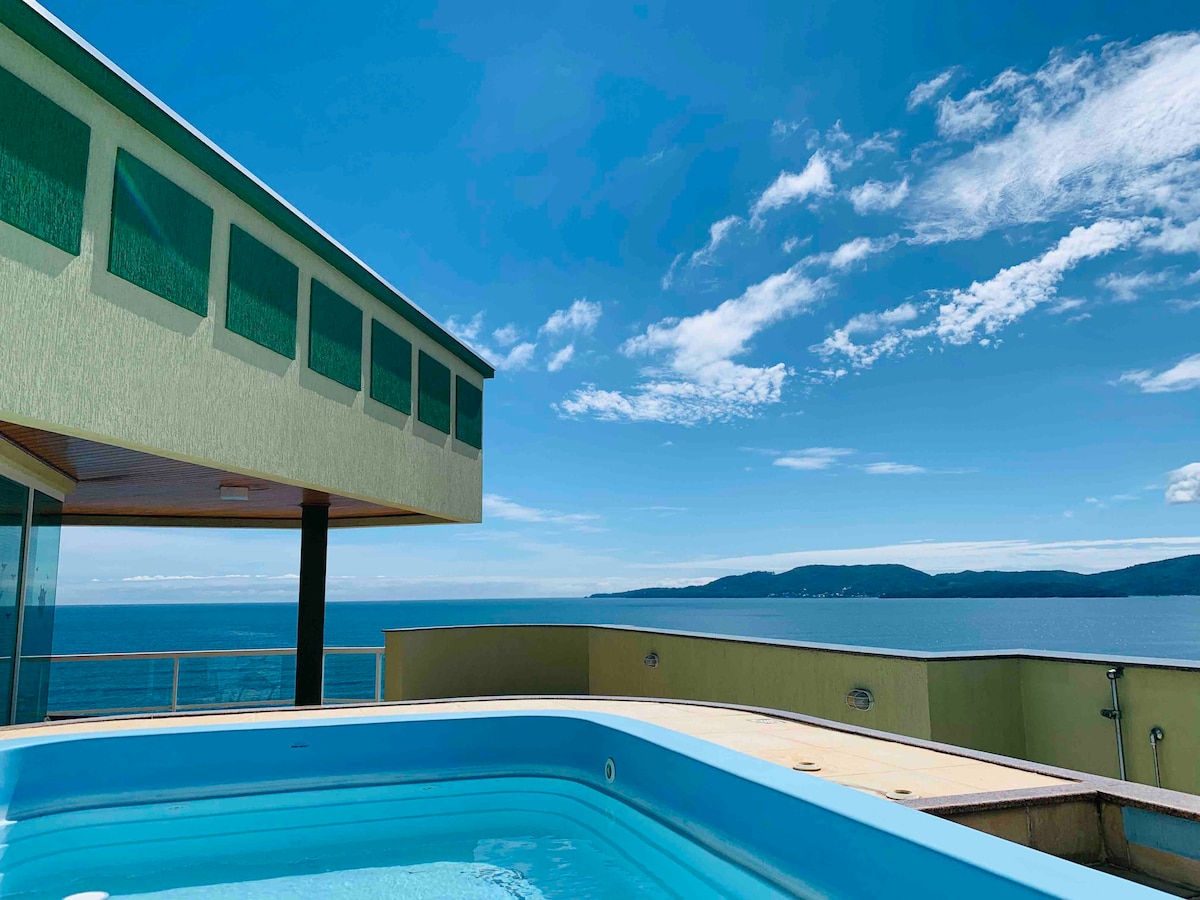 A private swimming pool is positioned on a spacious terrace, overlooking the expansive sea. Clear blue waters of the pool contrast with the blue sky, and the building is designed with large windows that frame the breathtaking coastal view.