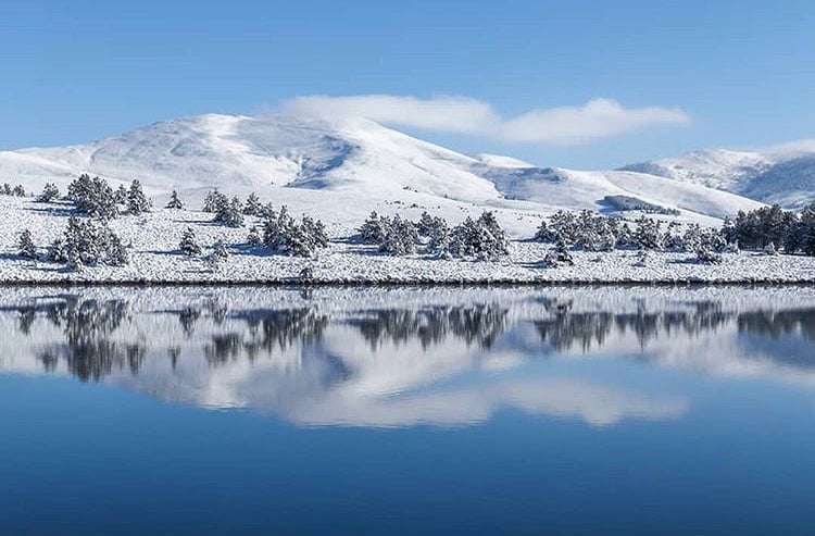 A serene winter landscape is presented, featuring snow-covered hills and trees mirrored in a calm, reflective body of water. The clear blue sky complements the tranquil scene, highlighting a peaceful atmosphere typical of a snowy retreat.