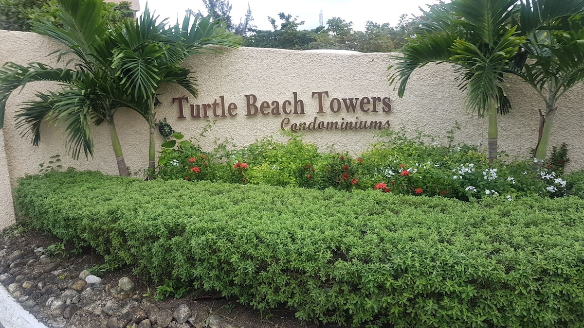 A landscaped entrance displays the 'Turtle Beach Towers' sign, surrounded by lush greenery. Vibrant flowers in shades of red and white are nestled among the neatly trimmed hedges, enhancing the inviting appearance of the condominium entrance.