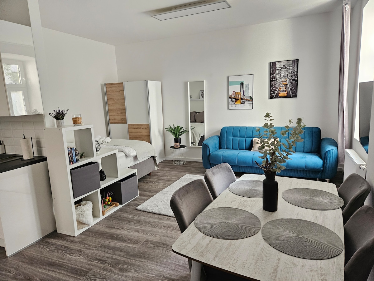 An open living area features a blue sofa, dining table, and light-colored decor. The kitchen provides modern appliances, while a cozy bed area is partially visible. The room is brightened by natural light through the window, enhancing the inviting ambiance.