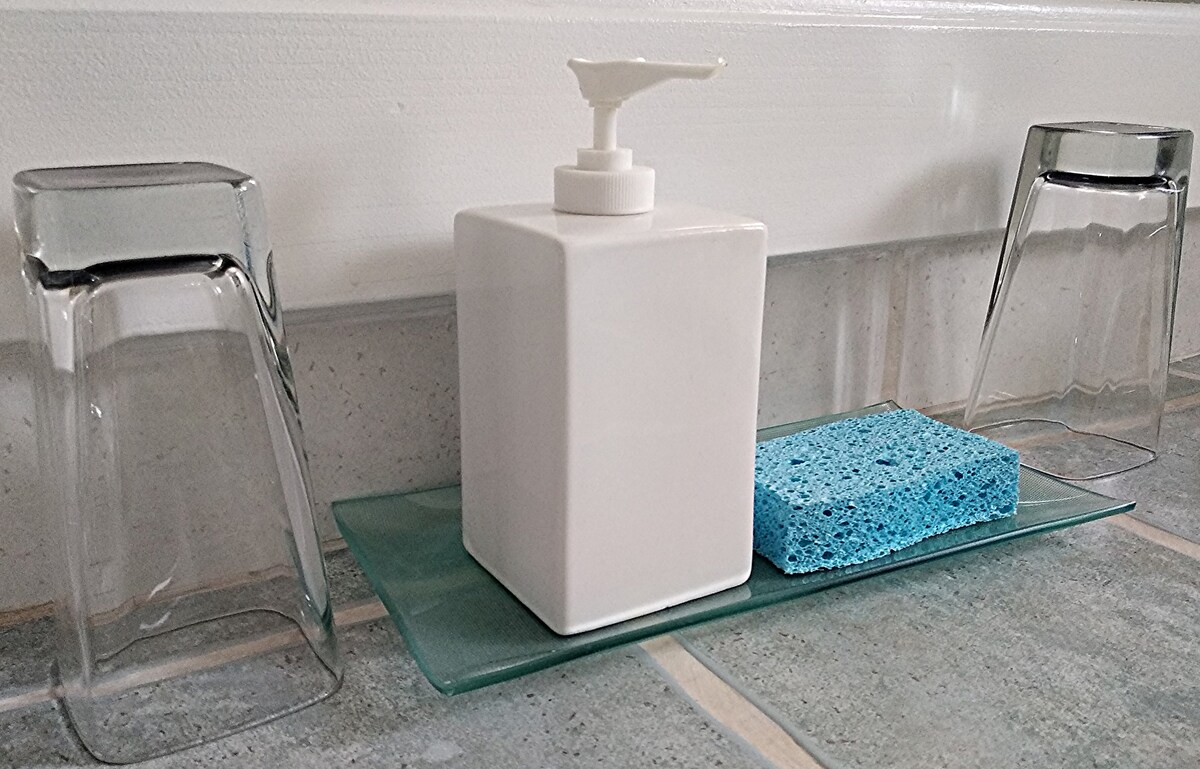 A clean countertop is displayed with a white soap dispenser positioned centrally on a glass tray. Two clear drinking glasses are placed upside down on either side, along with a blue sponge offering a soft texture contrast.