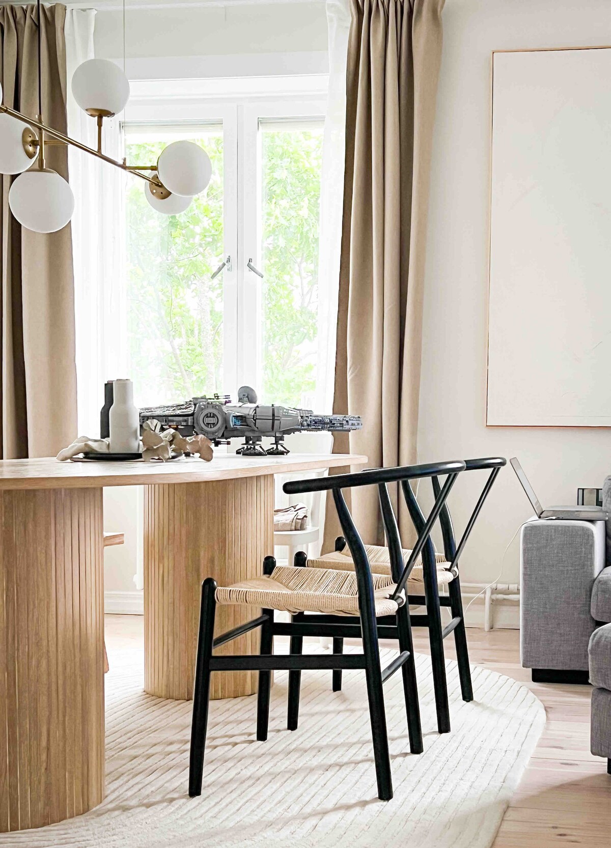 A dining area is presented with a round wooden table surrounded by black chairs featuring woven seats. Nearby, a large window is framed by light curtains, allowing natural light to illuminate the space. A decorative object sits on the table, adding interest to the setting.