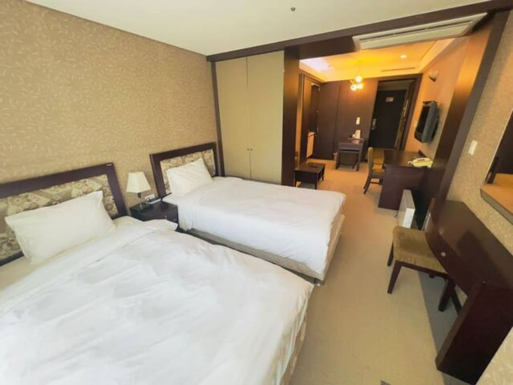 A spacious room features two super single beds with white linens. A wooden desk and chair are positioned near a mirror. Soft lighting enhances the warm tones of the wallpaper and carpet, creating a calm atmosphere. A closet is visible along one wall.