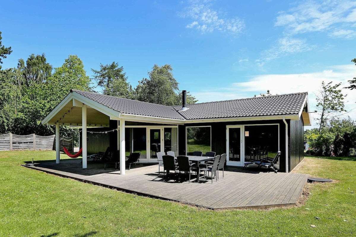 The exterior of the house is showcased with a spacious wooden deck, furnished with black outdoor chairs and a dining table. A hammock is suspended nearby, and large glass doors offer views into the living areas. Lush greenery surrounds the property under a clear blue sky.