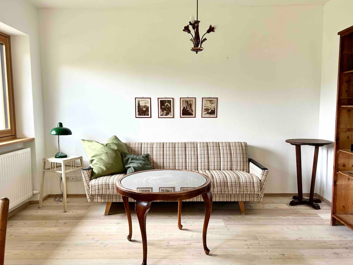 A compact living space is filled with vintage furnishings. A striped sofa sits across from a round wooden table, while a small green lamp adds a pop of color. Framed vintage photos adorn the wall, complemented by light streaming in from a window.