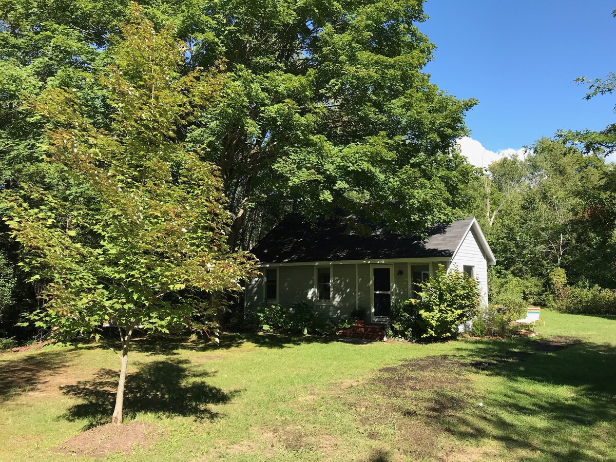 A cozy cottage is nestled among lush trees in a green, open space. The sun shines brightly on the well-maintained lawn, and a pathway leads to the entrance. The building features a welcoming facade with a sloped roof and a front porch surrounded by shrubs.