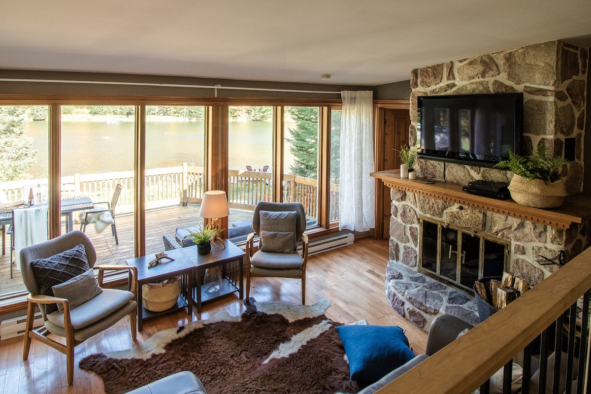 A comfortable seating area features two armchairs and a coffee table, situated beside a stone fireplace. Large windows offer a view of the tranquil lake outside, with a wooden deck visible in the background. Soft lighting enhances the welcoming atmosphere of the space.