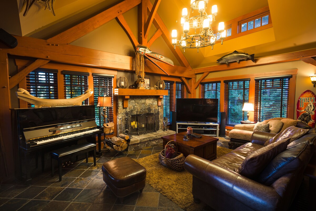 A spacious living area is highlighted by rich wooden beams and a stone fireplace. Comfortable leather seating surrounds a rustic coffee table. A grand piano is positioned nearby, and soft lighting from a chandelier enhances the warm ambiance. Blinds on the windows provide adjustable light control.
