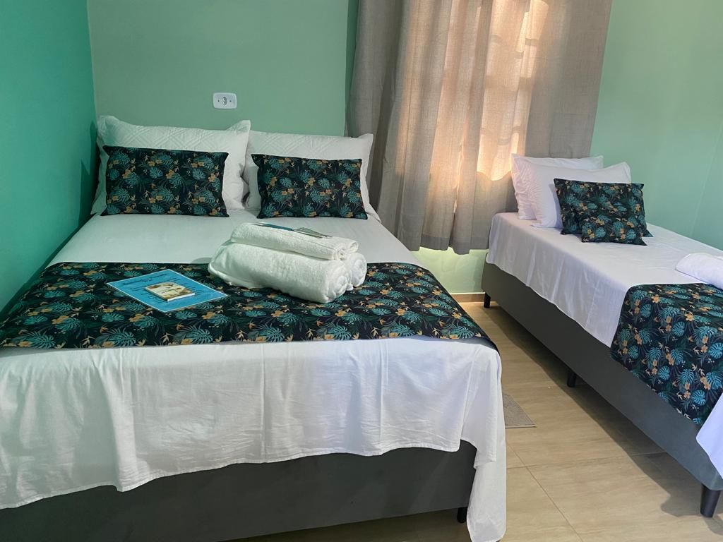 A sleeping area features two beds, each topped with patterned bedspreads. Towels are neatly arranged on the larger bed. Natural light filters through a window, partially covered by light curtains, illuminating the soothing green walls.