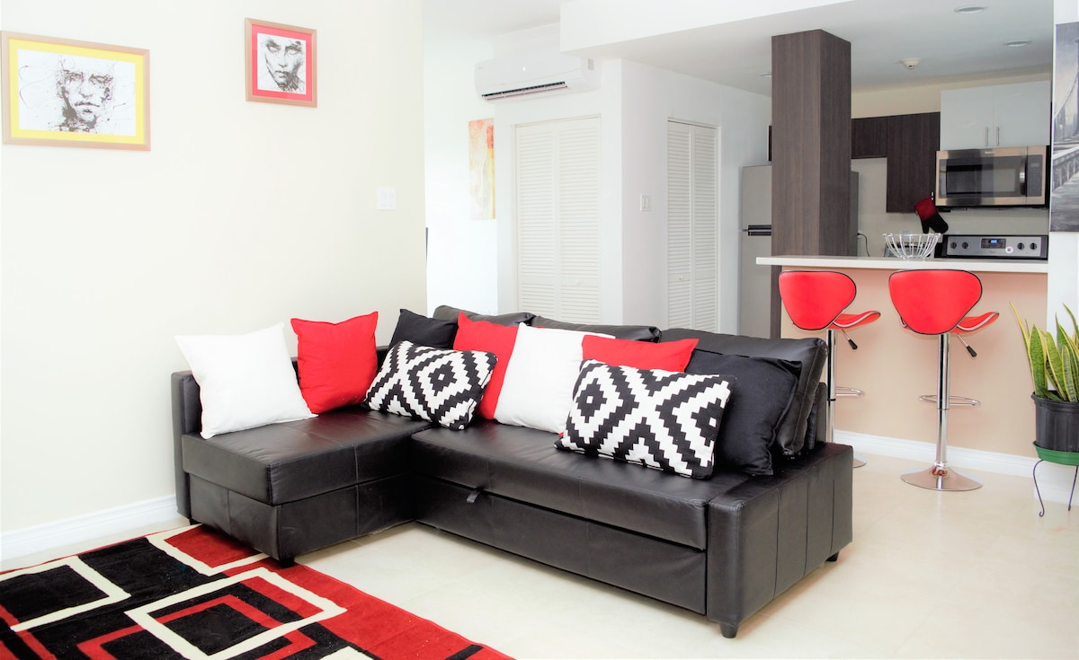 A black sectional sofa adorned with patterned cushions in black, white, and red is positioned in a bright living area. A stylish area rug adds a pop of color underfoot. In the background, kitchen bar stools in red complement the modern kitchen layout.