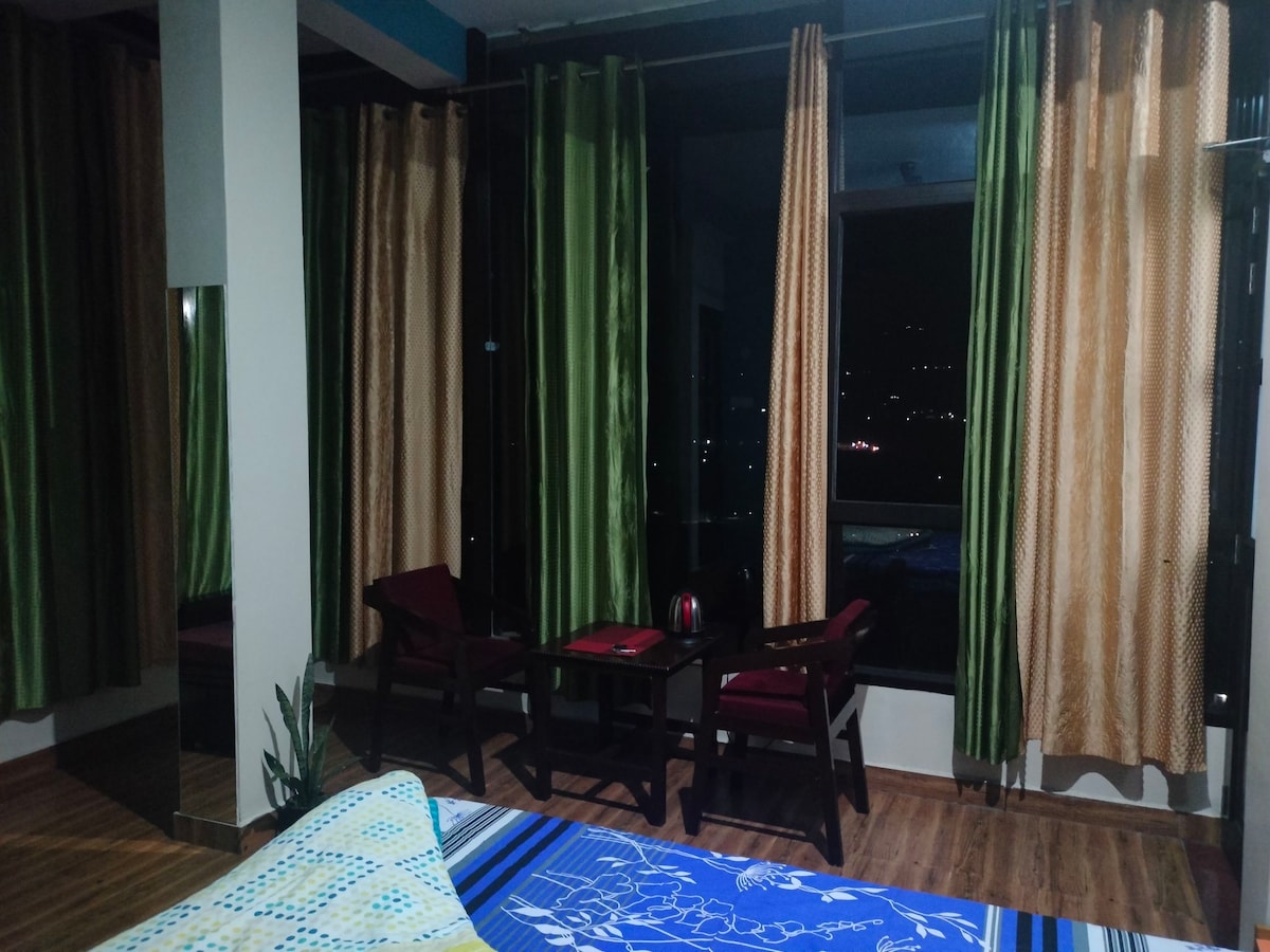 A cozy corner featuring two wooden chairs accompanied by a small table, positioned near large windows dressed in green and gold curtains. A vibrant blue bedspread lies on the spacious bed, while warm lighting enhances the inviting atmosphere of the room.