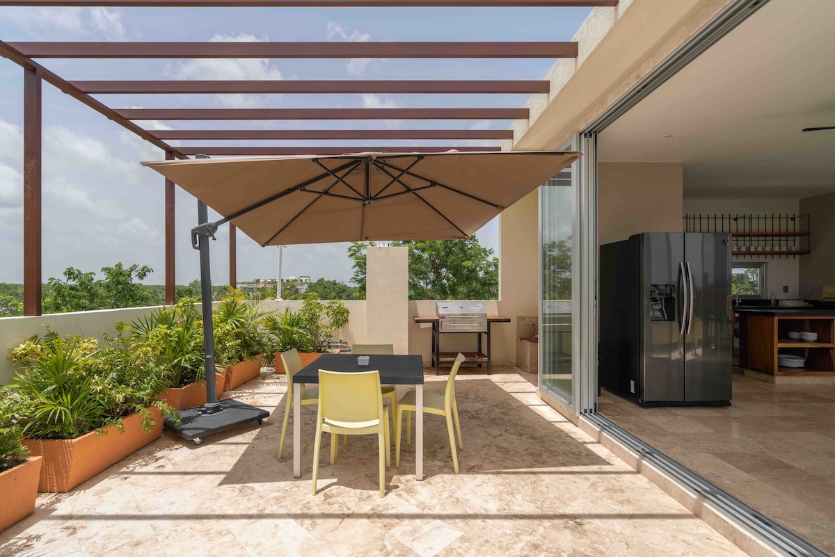 A spacious rooftop terrace is shown, featuring a large shaded umbrella over a black dining table accompanied by light-colored chairs. Green plants line the edges, providing a natural ambiance. A grill is positioned to one side, complementing an open and airy atmosphere.