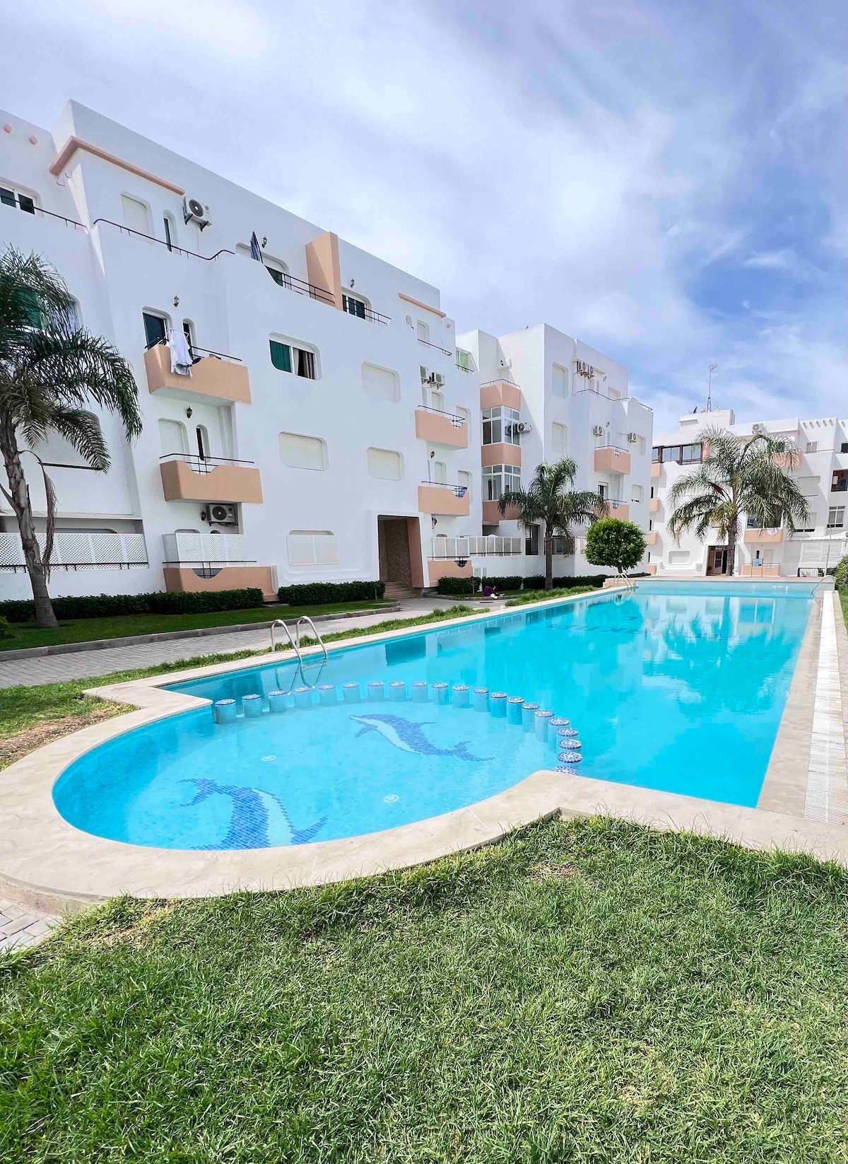 A clear blue swimming pool is bordered by well-maintained grass and a paved path. The surrounding white buildings are accented by palm trees, contributing to a serene outdoor atmosphere. The pool features a gentle curve and steps leading into the water.