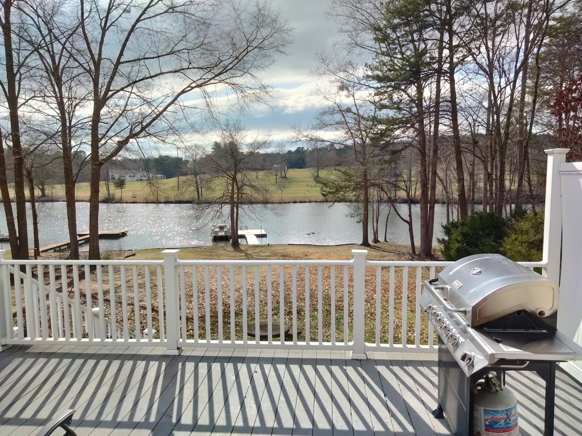 A spacious deck is shown overlooking a serene lake, with a barbecue grill positioned to one side. The surrounding area is framed by leafless trees and grassy banks, while the water reflects sunlight under a partly cloudy sky.
