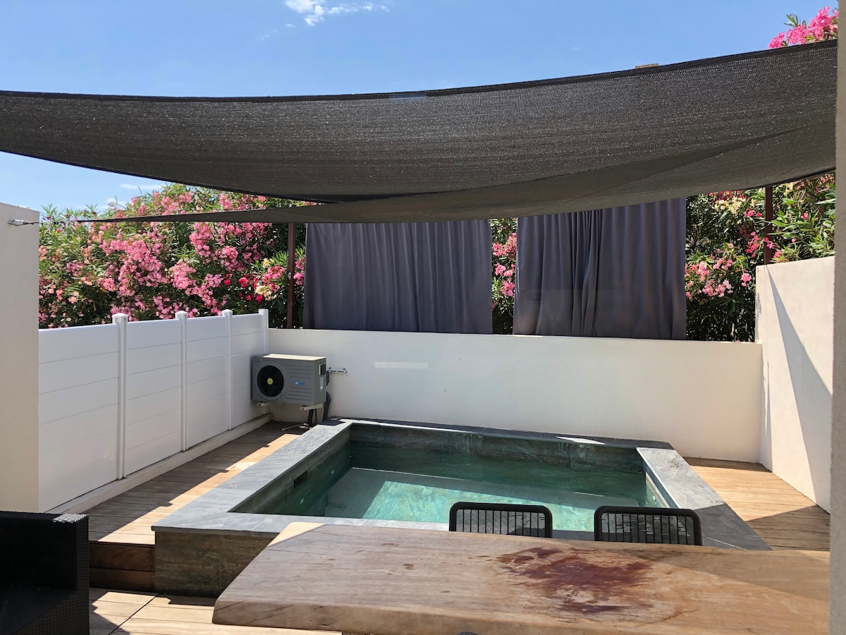 A private outdoor space features a small pool surrounded by wooden decking. Shade is provided by a dark canopy overhead. Lush flowering plants are visible in the background, adding color to the serene setting. Seating options include two black chairs near a wooden table.