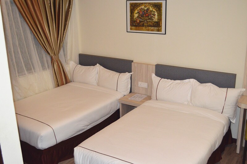 A neatly arranged room featuring two spacious beds with white linens and headboards. A wooden bedside table sits between the beds, and decorative curtains frame the window, allowing natural light to enter. A framed piece of art adorns the wall above the beds.