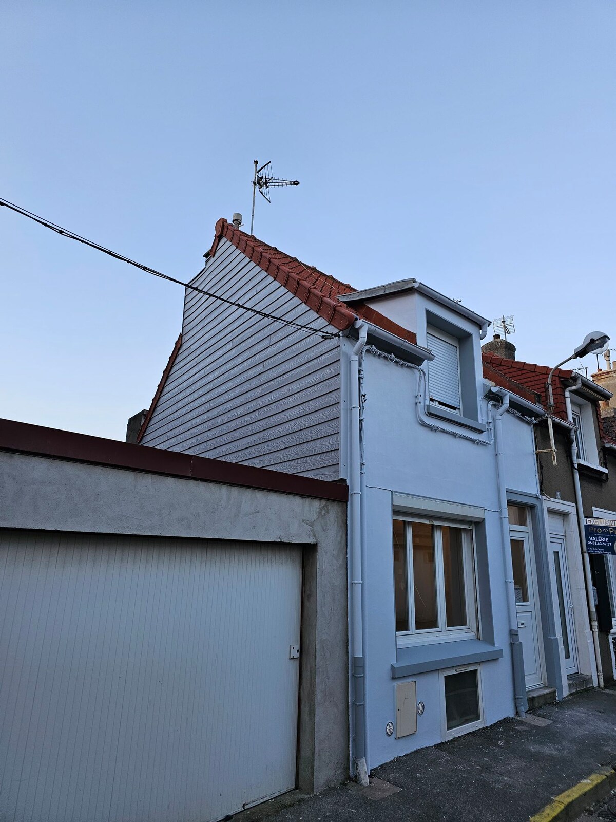 A two-story house is shown with light blue siding and a red-tiled roof. A small weather vane is visible on top, and a garage door is seen to one side. The structure is situated in a quiet area, with a clear blue sky overhead.