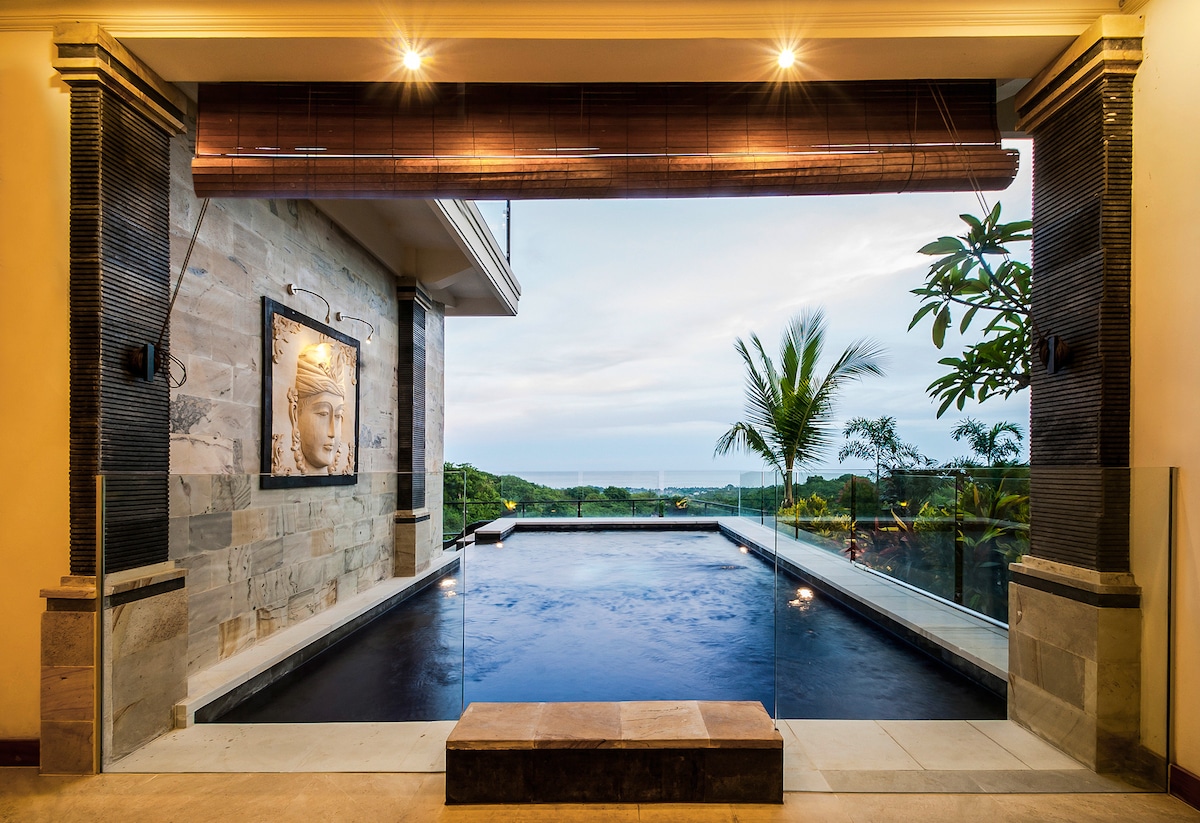 An infinity pool overlooks a scenic view, framed by large glass doors that lead to an outdoor area. Tropical plants are visible in the background, and warm lighting enhances the ambiance. A stone bench is positioned at the pool's edge.