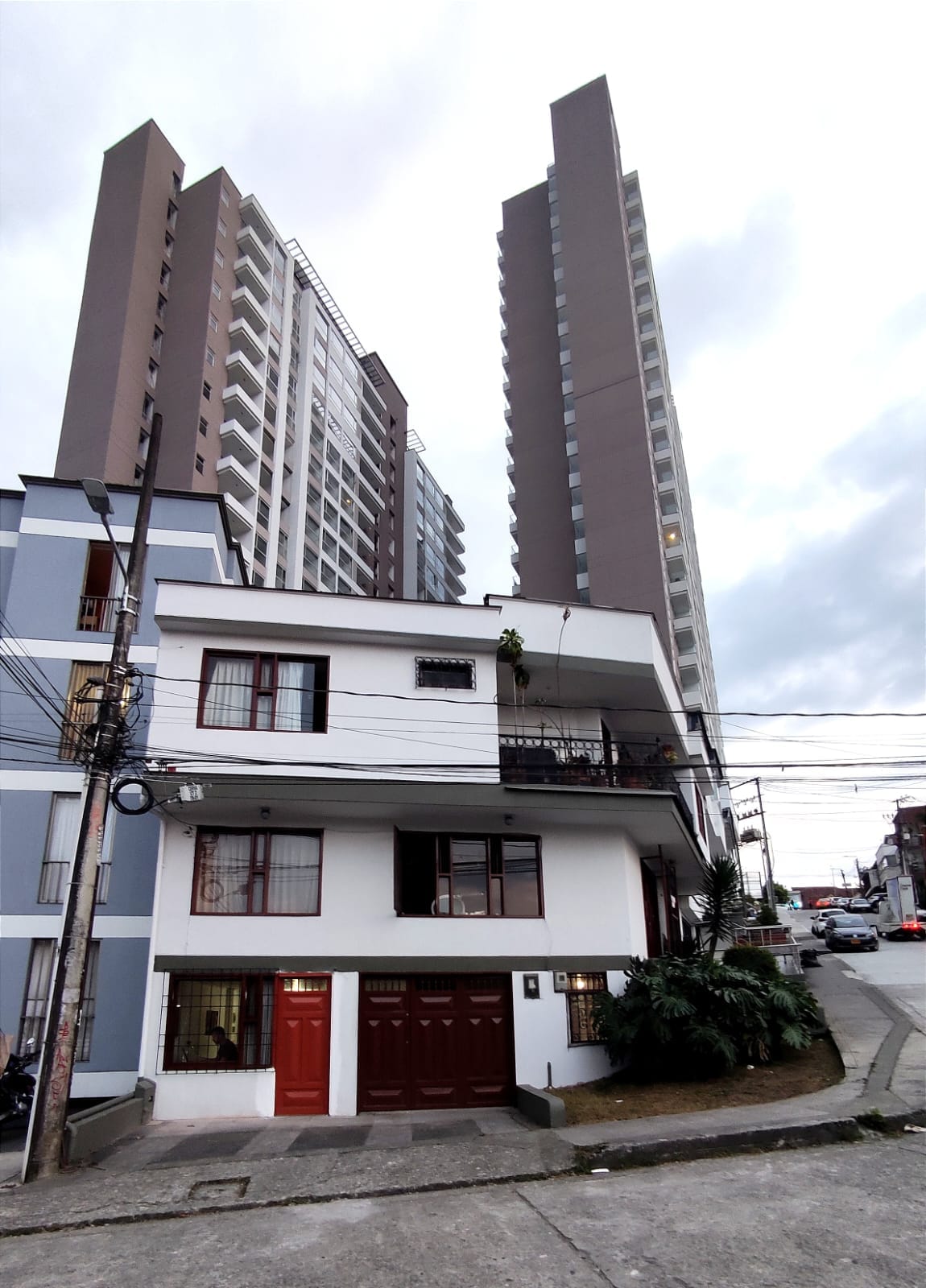 The image features a two-story white building with a red door, situated at the base of taller residential towers. Windows are visible along the facade, and greenery is present at the entrance. Power lines run overhead, indicative of an urban setting.