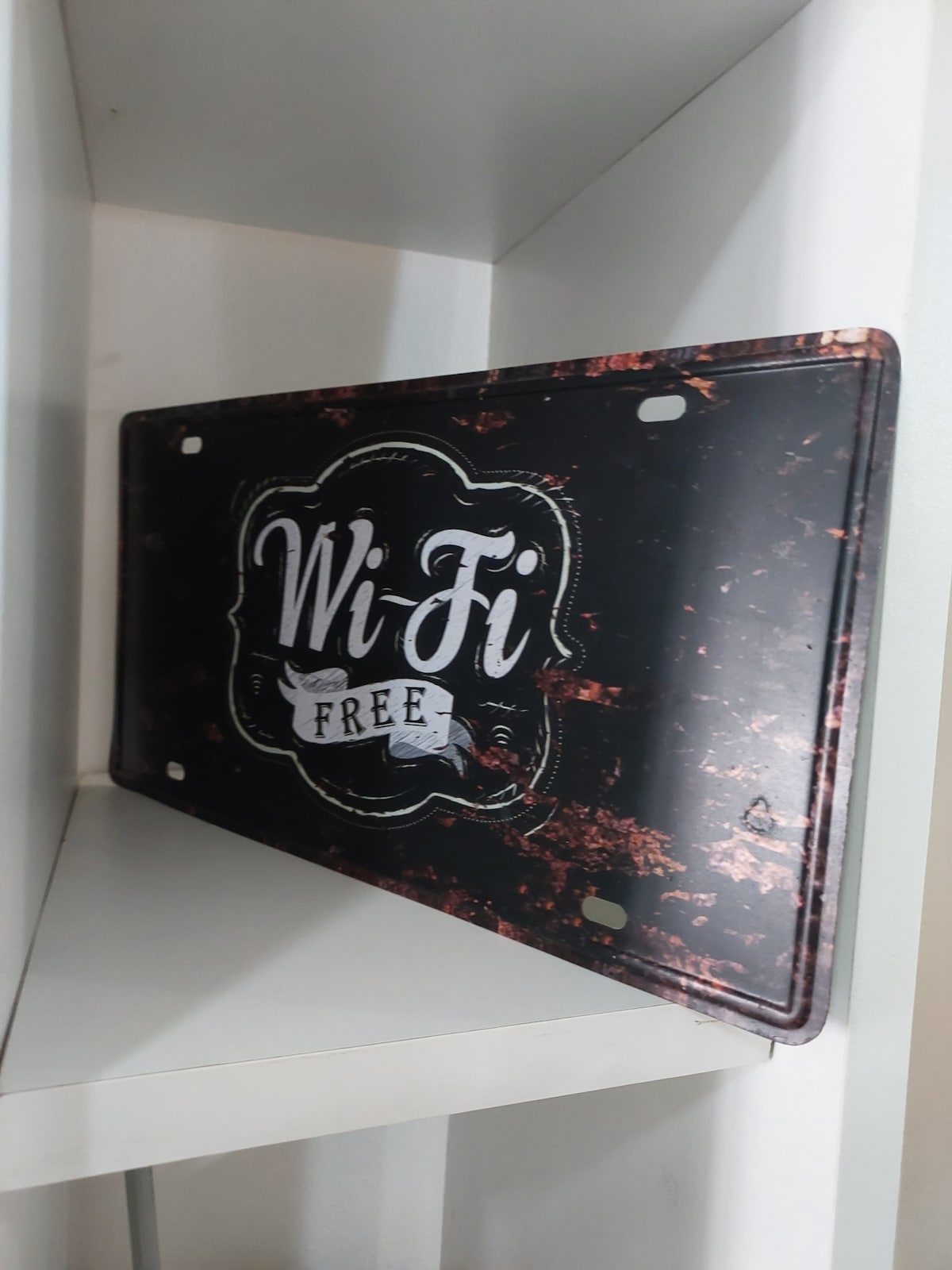 A decorative sign indicating 'Wi-Fi Free' is displayed on a shelf. The sign features a vintage design with a black background and white lettering, complemented by a subtle distressed finish that enhances its character.