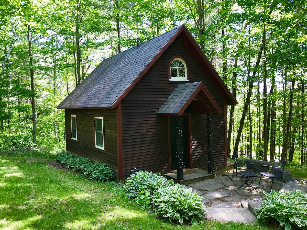 The Schoolhouse Cottage is a charming wooden structure nestled among tall trees, featuring a sloped roof and a welcoming front entrance. A patio with outdoor furniture is located on the stone pathway leading to the entrance, providing a peaceful setting surrounded by lush greenery.