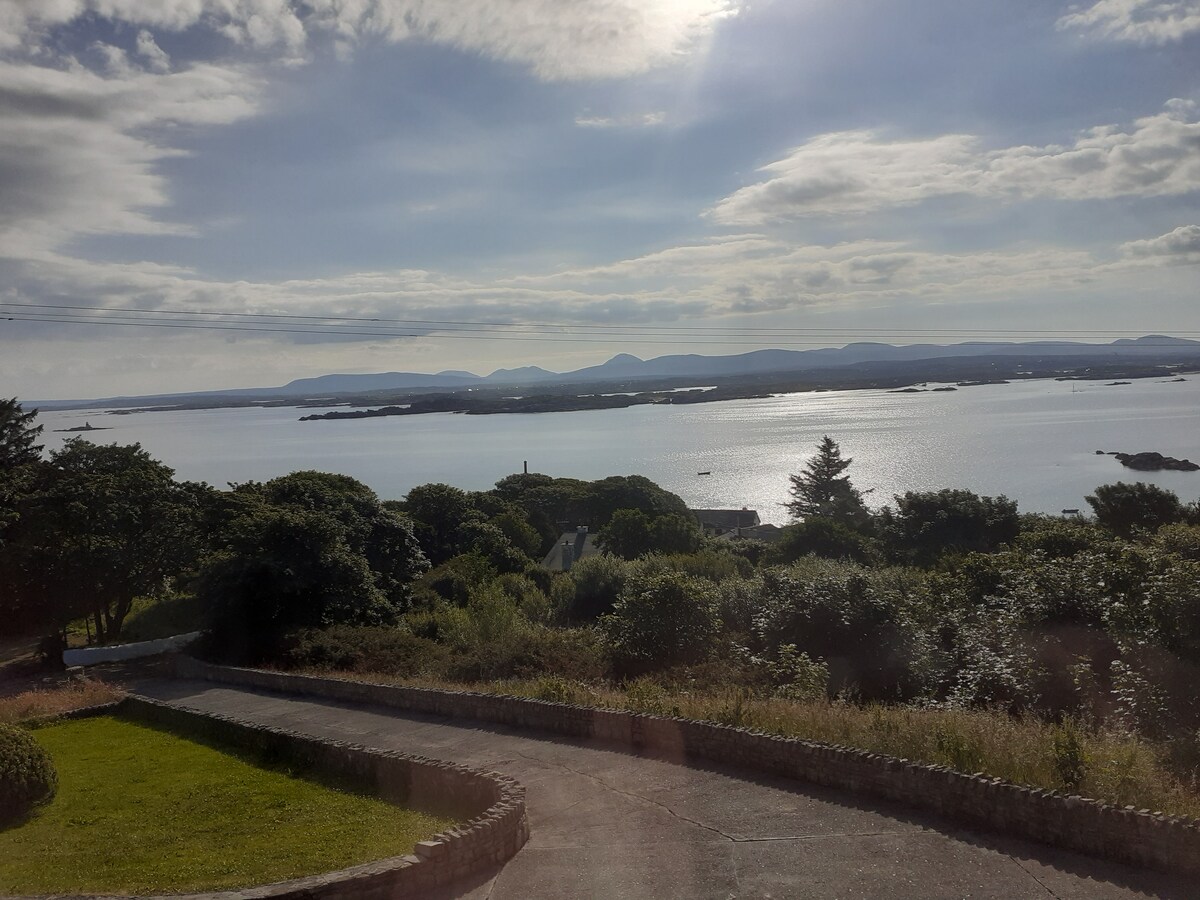 A panoramic view reveals tranquil waters stretching towards the horizon, with distant mountains softly outlined under a cloud-draped sky. Lush greenery frames the foreground, creating a serene atmosphere. The sunlight reflects off the water, enhancing the landscape's natural beauty.