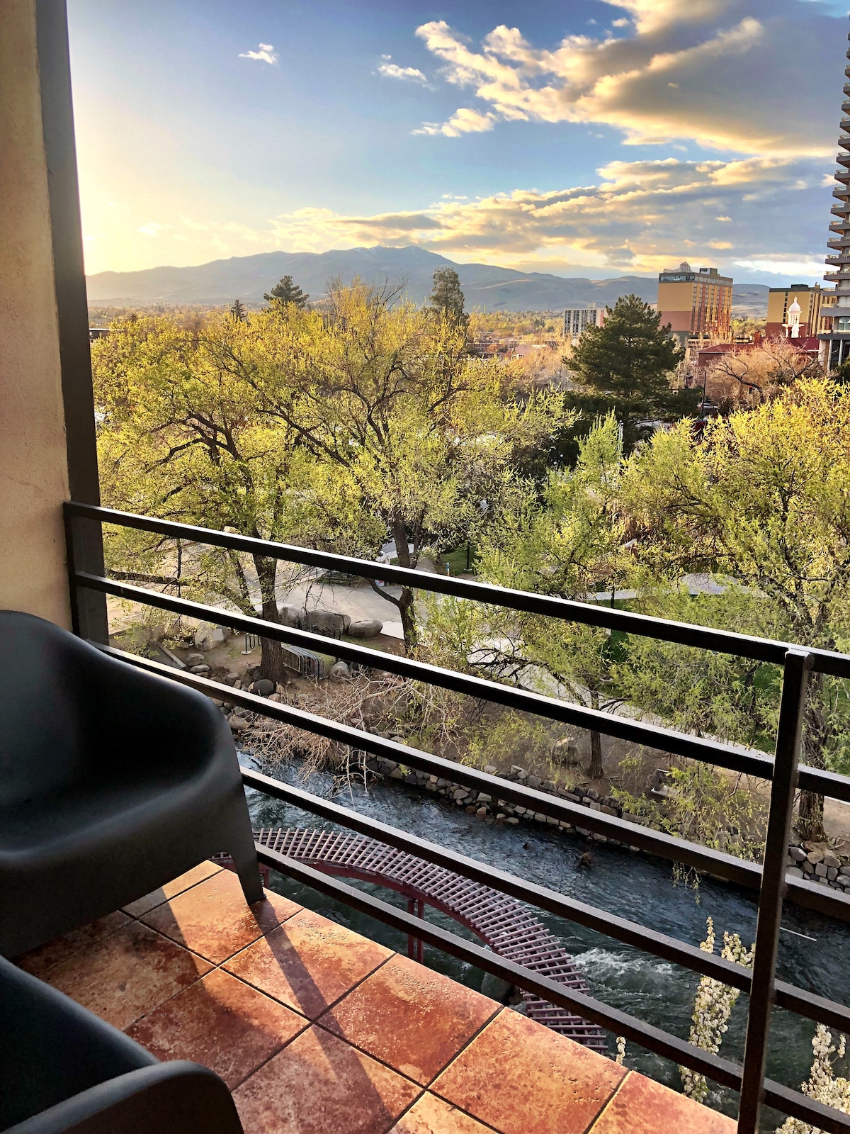 A balcony overlooks a serene river lined with lush trees. Comfortable seating is positioned on the tiled surface, with distant mountains and a city skyline visible in the background. The sky is partially cloudy, capturing the soft light of sunset.