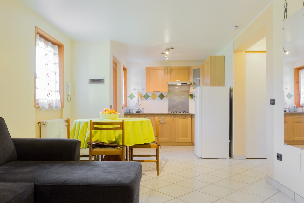 The kitchen area features light wood cabinetry and modern appliances, including a refrigerator and gas stove. A dining table with yellow tablecloth is set for meals. Soft lighting enhances the warm ambiance in the spacious layout, which is complemented by tiled flooring.