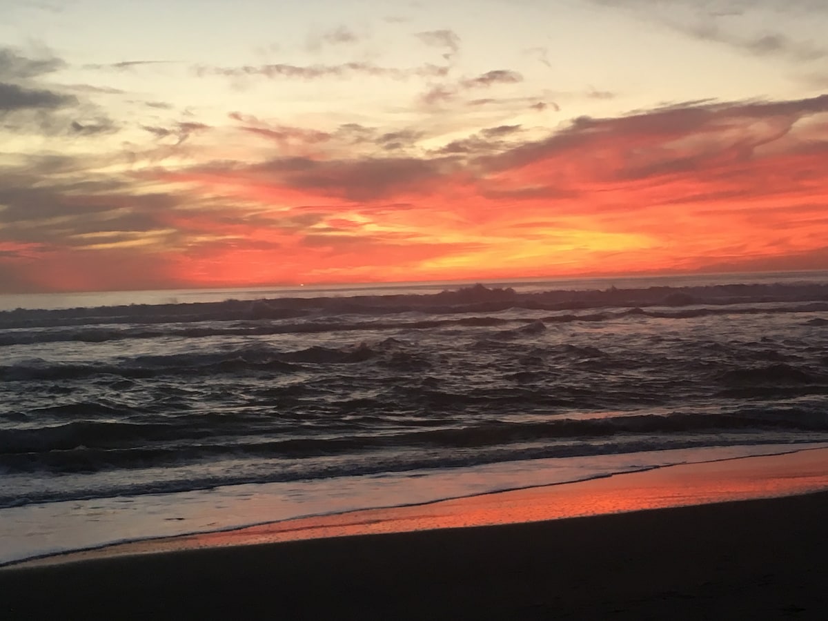 A vibrant sunset paints the sky in shades of orange and pink, reflecting on the wet sand of the beach. Waves crash gently in the foreground, while a cloudy sky adds depth to the serene coastal atmosphere.