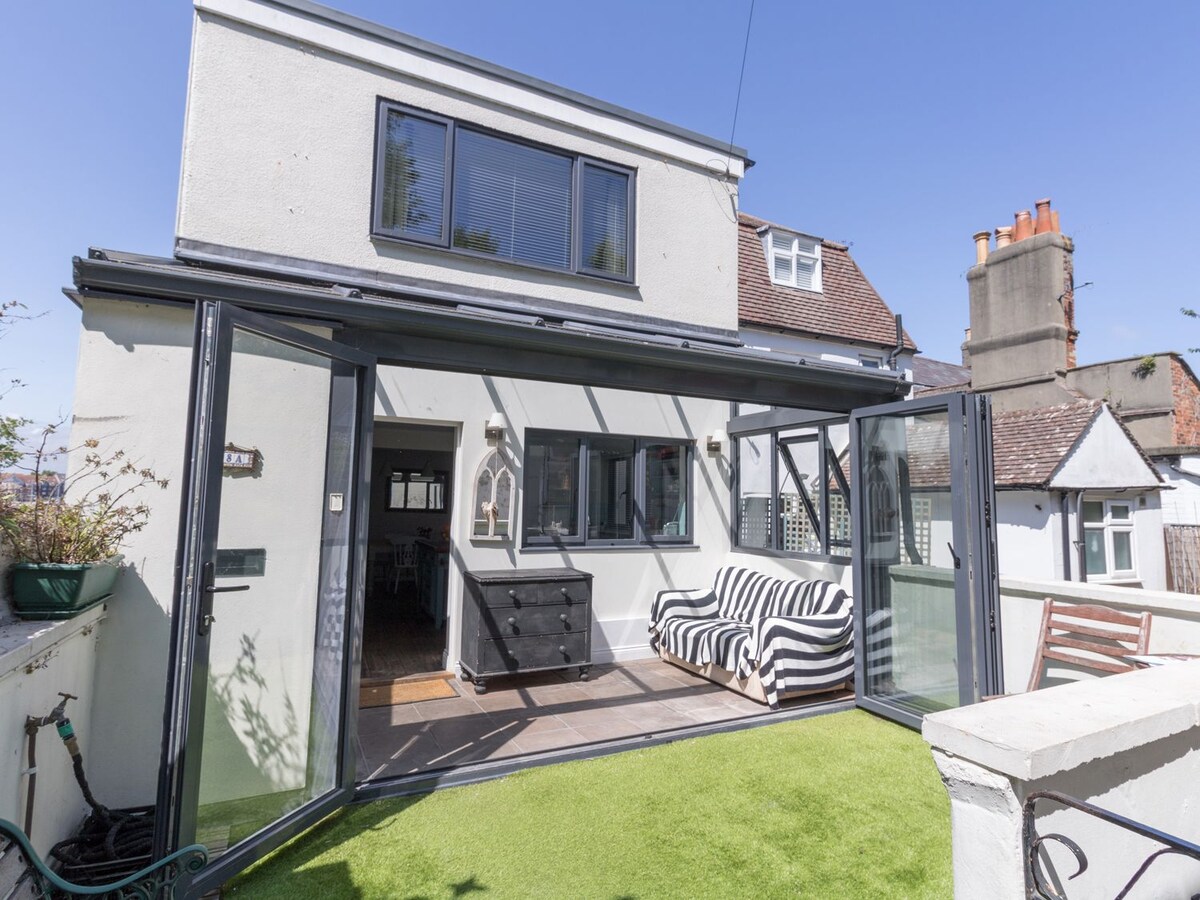 A conservatory features tri-fold doors that open to a walled garden, showcasing artificial grass. Inside, a double sofa and armchair are positioned for relaxation. Natural light floods the space, highlighting the modern decor and creating an inviting atmosphere for gatherings.
