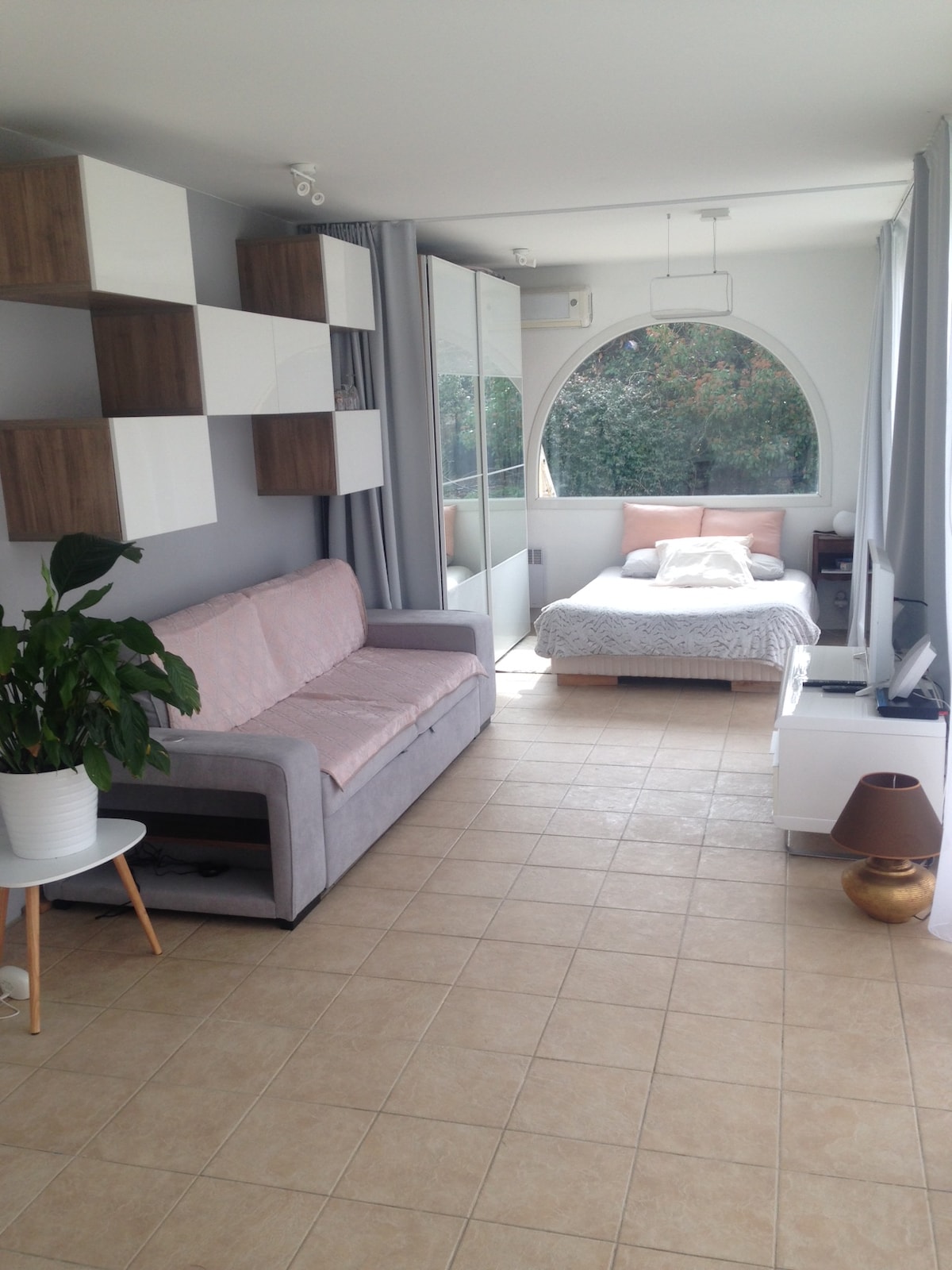 A bright living space features a comfortable sofa alongside a small table with a potted plant. An arched window offers views of greenery outside. The area includes a cozy sleeping arrangement with light bedding, complemented by soft hues throughout the room.