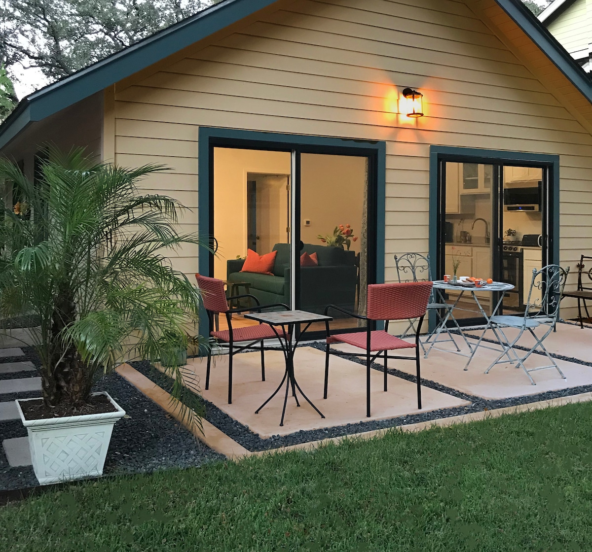 An outdoor patio is depicted, featuring two tables with chairs and a potted palm. Large sliding doors provide a view of the interior, which includes a sofa and kitchen area. The surrounding lawn is neatly maintained, enhancing the inviting space.