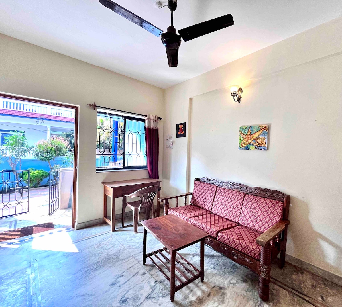 A well-lit living area features a wooden sofa with a patterned fabric, accompanied by a simple wooden coffee table. A small desk and chair provide a cozy workspace. Natural light streams through the window, with a view of the surrounding garden area, enhancing the room’s inviting nature.