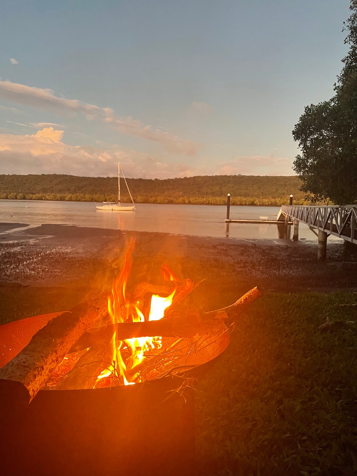 A fire pit set on a grassy area is shown, with a warm flame crackling. In the background, a calm waterway reflects soft light from the setting sun, while a sailboat is anchored near a wooden jetty surrounded by lush greenery.