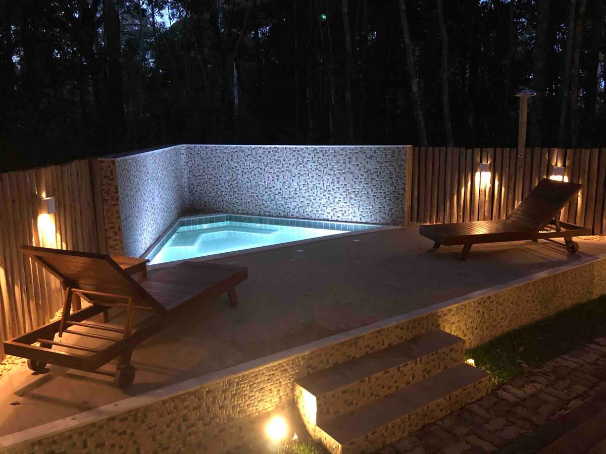 An illuminated private pool area is presented, showcasing a shallow pool bordered by natural stone. Two wooden lounge chairs are positioned nearby, allowing for relaxation. The surrounding environment includes tall trees, creating a serene and secluded atmosphere.