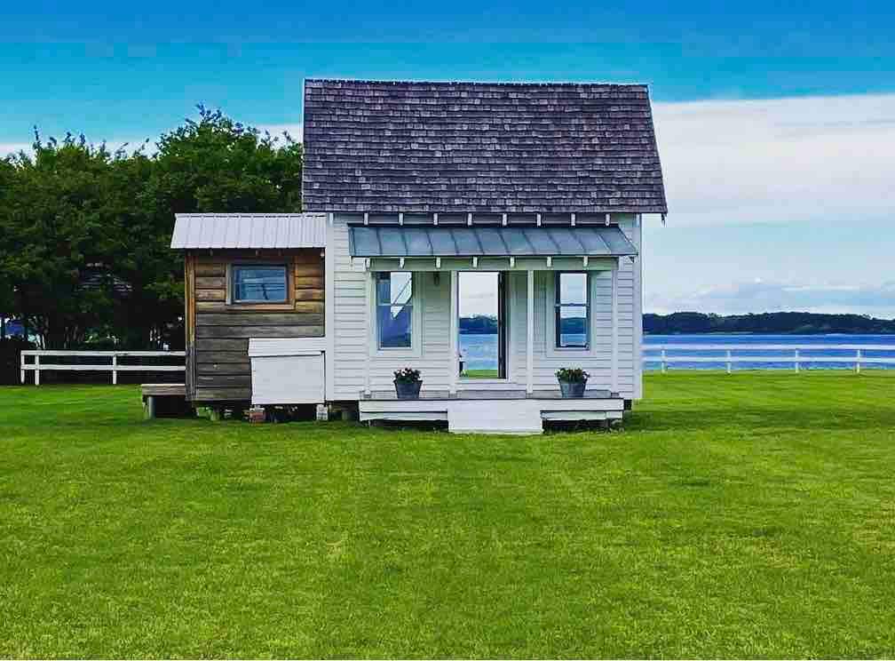 A charming two-story cottage is set on a lush green lawn, with a clear view of the Chesapeake Bay in the background. The cottage features large windows and a sloped roof, with a small wooden addition on one side, surrounded by well-maintained grass.